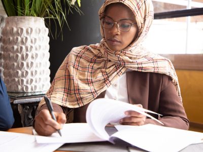 Muslim African American businesswoman scanning contract. Business women wearing hijabs, sitting in cafe and reading document with pen. Signing agreement concept